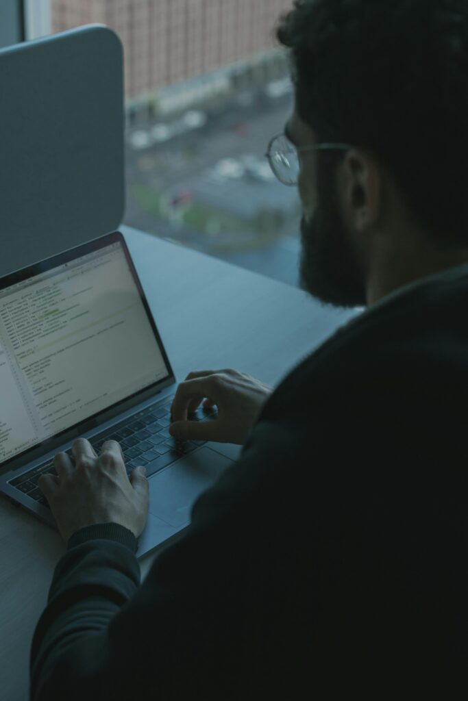 A man coding on his laptop by a window in an office setting, showcasing technology work.